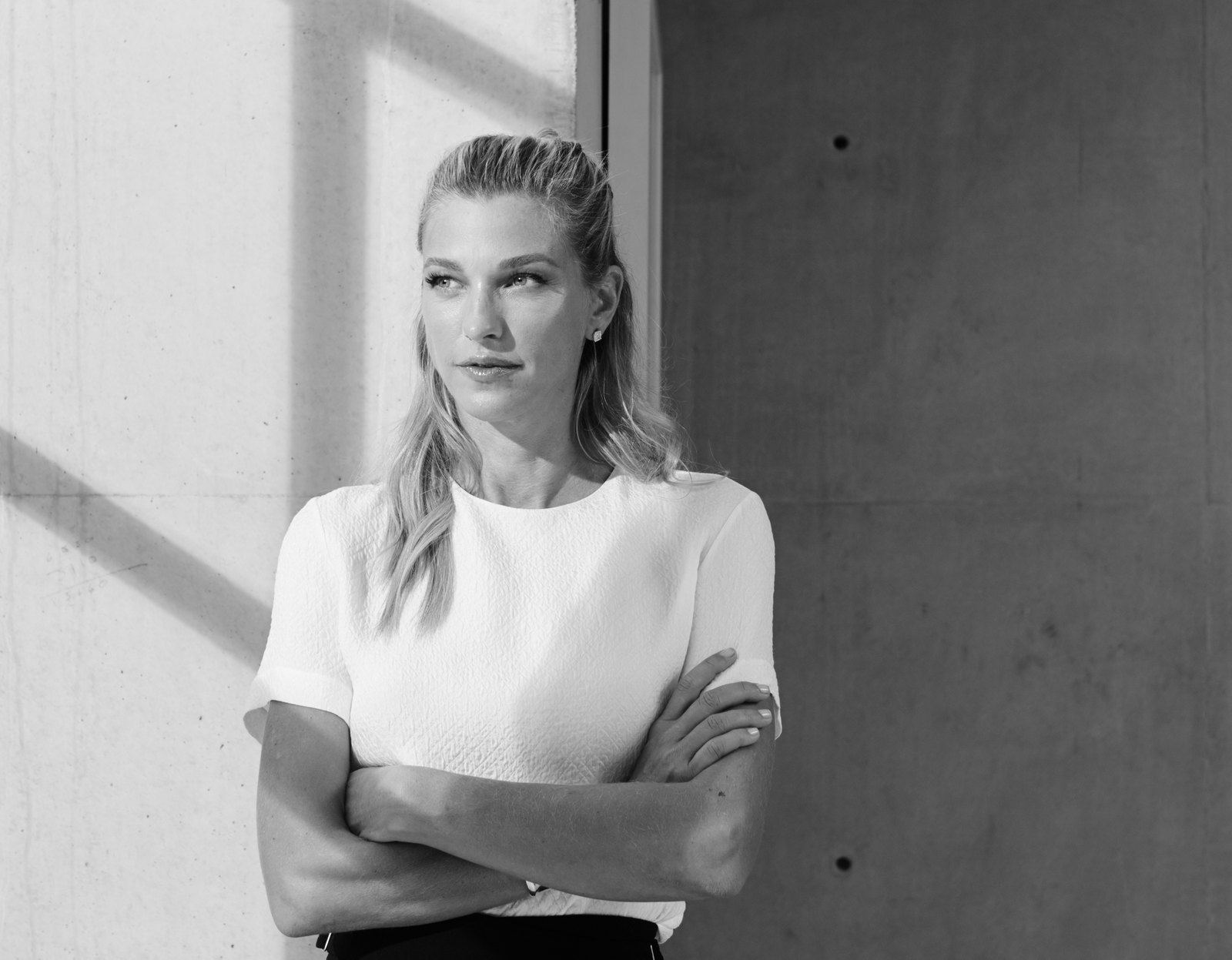 Nicole Junkerman standing by a sunlit concrete wall, arms folded, looking calmly ahead