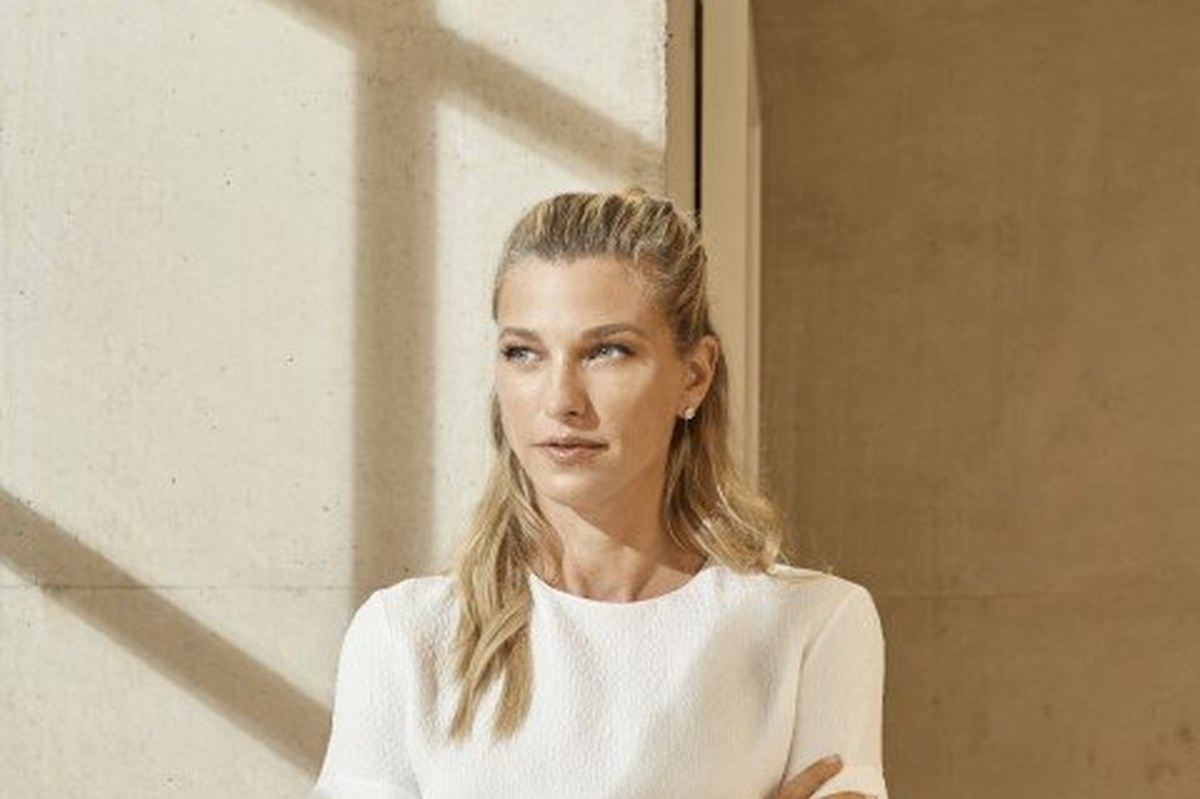 Nicole Junkerman in a white top with her arms folded, standing in bright daylight beside a pale wall